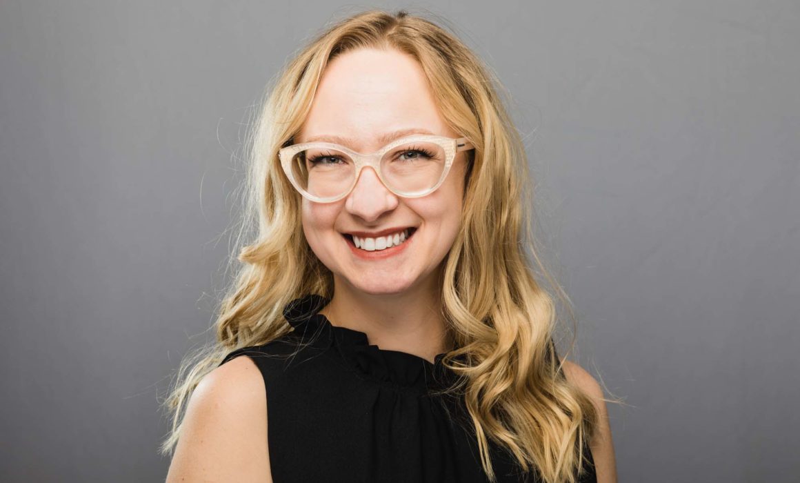headshot of female professional in black shirt wearing glasses, set against a neutral gray backdrop